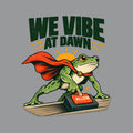 Illustration of a green frog in a red cape pressing a glowing red "ALLOW" button on a small desktop controller, with a rising sun behind it and the bold headline "WE VIBE AT DAWN" above, on a plain cream background. Artwork shown on the Sport Grey garment colour.