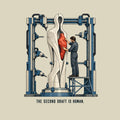 Illustration of a tall pale machine-milled humanoid sculpture with a central cavity, held in an industrial gantry; a worker on a platform installs a rough, hand-carved red heart-shaped piece into the opening. Caption reads: THE SECOND DRAFT IS HUMAN. Artwork shown on the Natural garment colour.