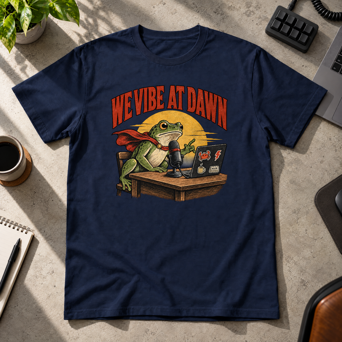 Illustrated frog wearing a red cape sits at a wooden desk before a microphone, one hand raised, beside a laptop with crab, lightning bolt, coffee mug and ALLOW stickers, under a yellow dawn sun and the arched red headline WE VIBE AT DAWN. Shown on the Navy tee.