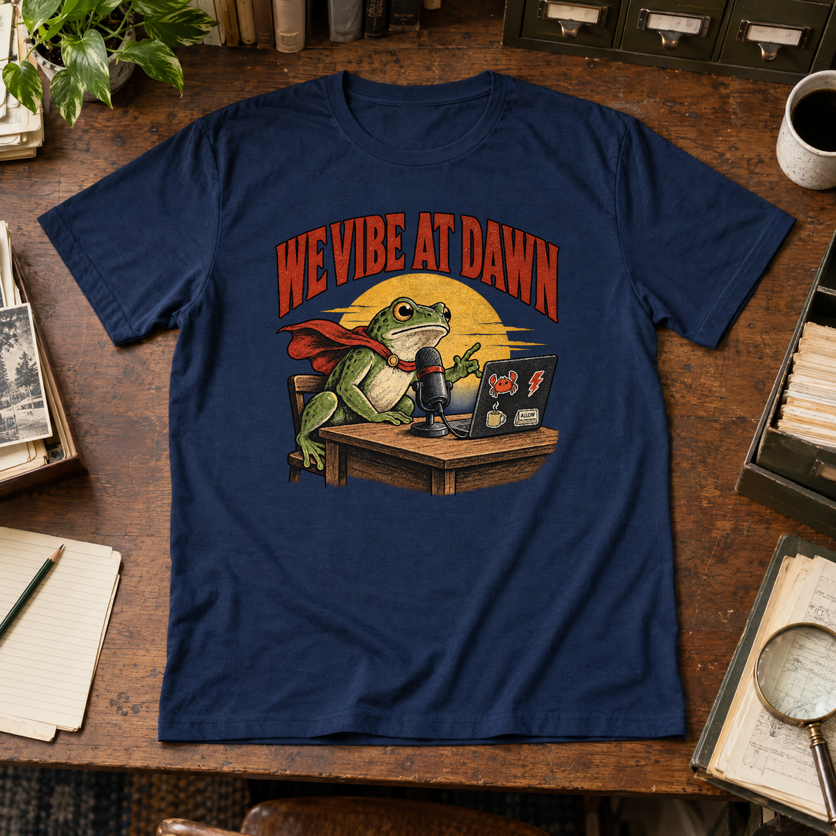 Illustrated frog wearing a red cape sits at a wooden desk before a microphone, one hand raised, beside a laptop with crab, lightning bolt, coffee mug and ALLOW stickers, under a yellow dawn sun and the arched red headline WE VIBE AT DAWN. Shown on the Navy tee.
