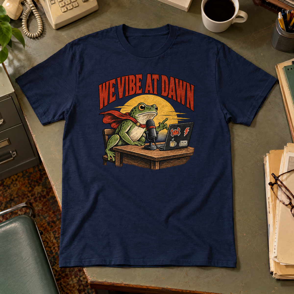 Illustrated frog wearing a red cape sits at a wooden desk before a microphone, one hand raised, beside a laptop with crab, lightning bolt, coffee mug and ALLOW stickers, under a yellow dawn sun and the arched red headline WE VIBE AT DAWN. Shown on the Navy tee.