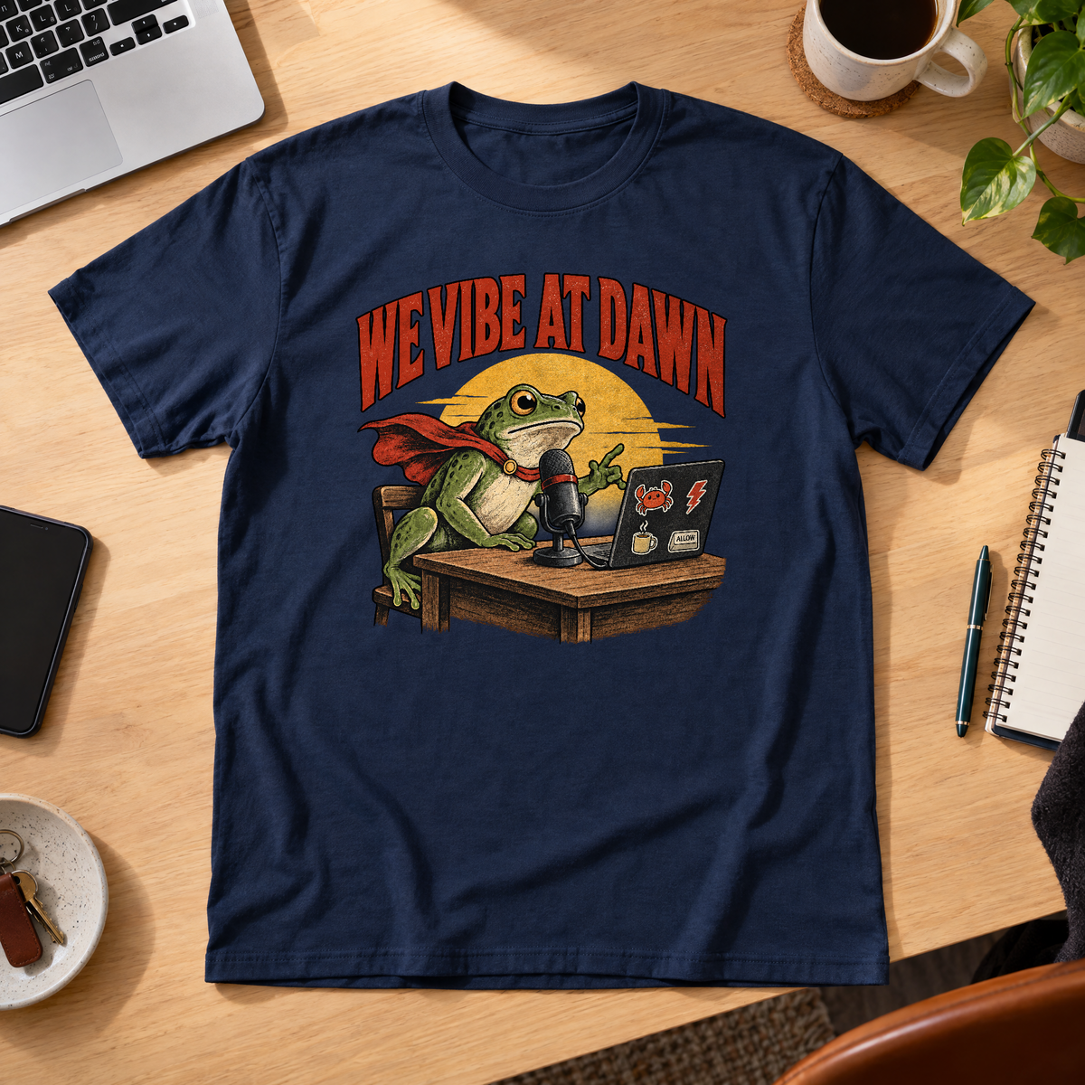 Illustrated frog wearing a red cape sits at a wooden desk before a microphone, one hand raised, beside a laptop with crab, lightning bolt, coffee mug and ALLOW stickers, under a yellow dawn sun and the arched red headline WE VIBE AT DAWN. Shown on the Navy tee.