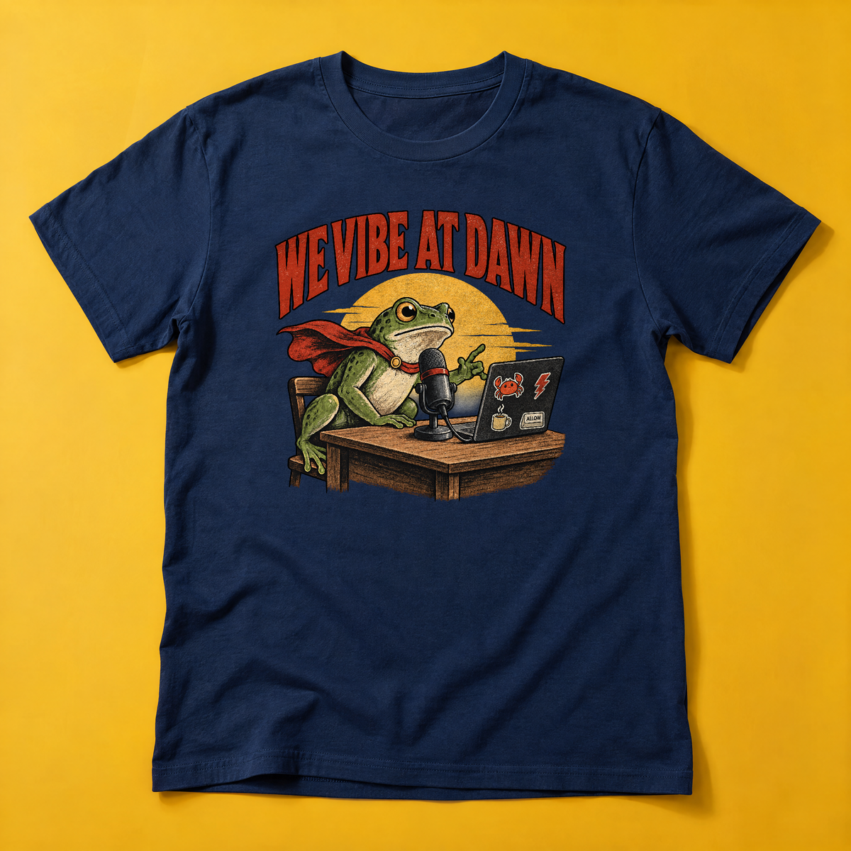 Illustrated frog wearing a red cape sits at a wooden desk before a microphone, one hand raised, beside a laptop with crab, lightning bolt, coffee mug and ALLOW stickers, under a yellow dawn sun and the arched red headline WE VIBE AT DAWN. Shown on the Navy tee.