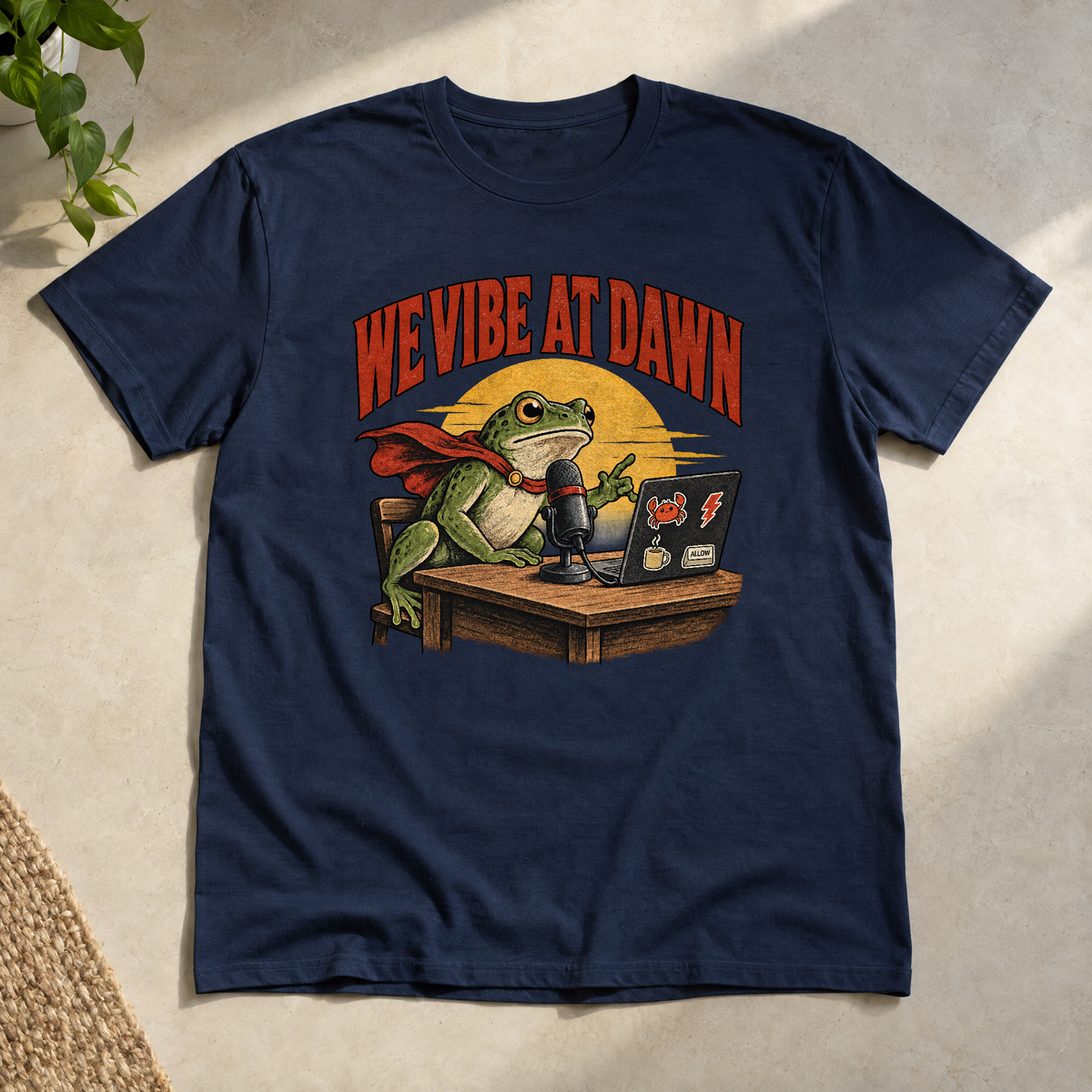 Illustrated frog wearing a red cape sits at a wooden desk before a microphone, one hand raised, beside a laptop with crab, lightning bolt, coffee mug and ALLOW stickers, under a yellow dawn sun and the arched red headline WE VIBE AT DAWN. Shown on the Navy tee.