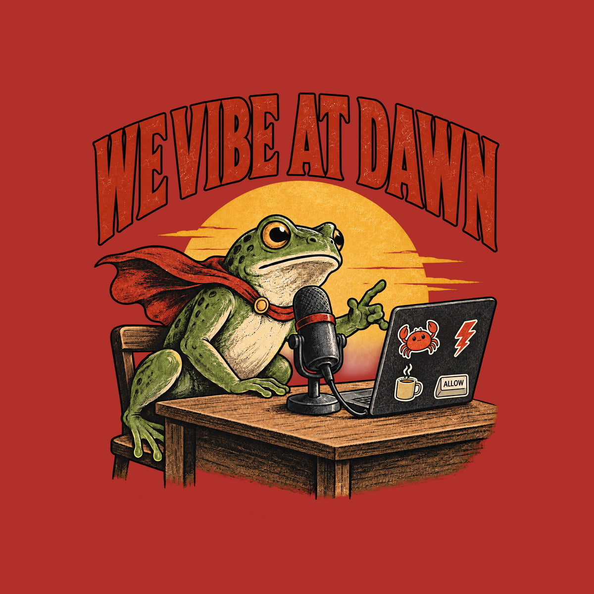 Illustrated frog wearing a red cape sits at a wooden desk before a microphone, one hand raised, beside a laptop with crab, lightning bolt, coffee mug and ALLOW stickers, under a yellow dawn sun and the arched red headline WE VIBE AT DAWN. Artwork shown on the Red garment colour.